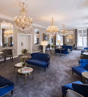 Elegant lounge med sittplatser i blå sammet, ljuskronor och utsmyckade speglar på vita väggar. Grå matta och runda bord med dekorativa växter förstärker den sofistikerade atmosfären. Stora fönster ger naturligt ljus.