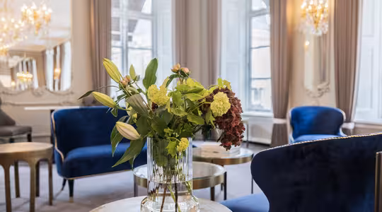 En lyxig lounge med blå sammetsstolar och runda träbord. En glasvas med diverse blommor sitter på ett bord i förgrunden. Eleganta ljuskronor och stora fönster med grå gardiner pryder det ljusa utrymmet.