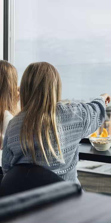 Två personer som sitter vid ett stort fönster på en restaurang och njuter av en skål med mat. En person pekar utåt och uppmärksammar stadsbilden med olika byggnader och en molnig himmel i bakgrunden.