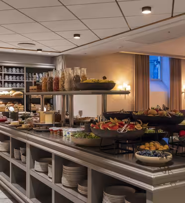 En väl upplyst hotellbuffé med olika matvaror utställda på en lång bänkskiva. Synliga rätter inkluderar frukt, grönsaker, flingor och kakor. Tallrikarna är prydligt staplade under disken, mot en bakgrund av eleganta gardiner och hyllor.