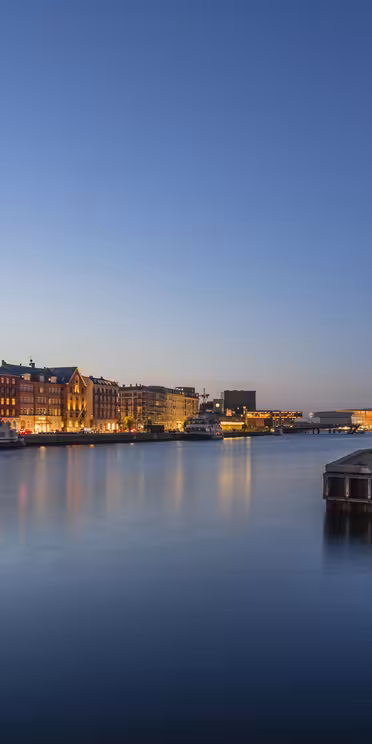 En lugn kvällsscen av en stadsbild vid vattnet. Upplysta byggnader kantar det lugna vattnet under en klarblå himmel som reflekterar levande ljus. En träbrygga sträcker sig in i floden, vilket ger djup till den fridfulla stadsvyn.
