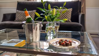 Ett glasbord rymmer en tallrik med chokladtäckta jordgubbar, två glas champagne, en ishink med en champagneflaska och en vas med liljor; en soffa med mönstrade kuddar syns i bakgrunden.