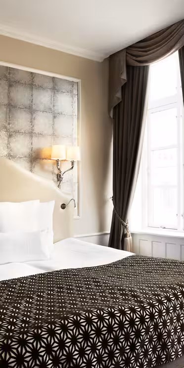 Ett väl upplyst hotellrum med en stor säng med vita kuddar och en svart mönstrad filt. Väggarna är ljusa med en dekorativ panel bakom sängen. Två eleganta vägglampor lyser upp området. Ett fönster med tunga gardiner finns till höger.
