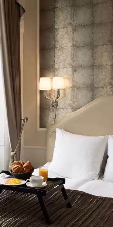 Ett mysigt hotellrum har en snyggt bäddad säng med vita kuddar och en beige sänggavel. En bricka med frukostartiklar, inklusive croissanter och juice, ligger på sängen. Två vägglampetter lyser upp utrymmet nära ett fönster med fördragna gardiner.