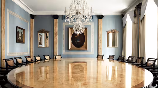 Elegant konferensrum med ett stort, polerat ovalt bord omgivet av svarta stolar. Rummet har ljusblå väggar, guldkantade speglar och en stor ljuskrona. Ett inramat porträtt hänger på den bortre väggen mellan höga fönster med draperier.